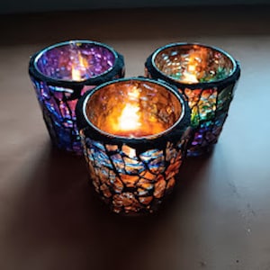May include: Three mosaic glass candle holders with lit candles inside. The holders have a black rim and a colorful, cracked glass design in shades of purple, orange, and green. The candles emit a warm, inviting glow.
