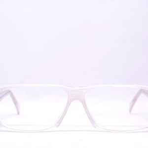 May include: A pair of spectacles featuring a translucent, pale pink frame with a subtle marbled texture. The rectangular lenses are clear, and the frame includes small, circular gold-toned metal accents at the hinges where the temples connect.