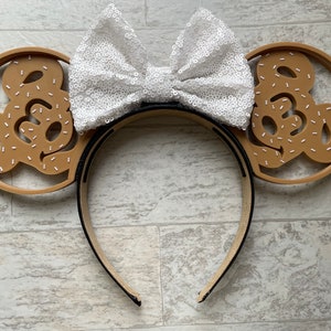 May include: A pair of brown mouse ears with a white bow. The ears are shaped like a Mickey Mouse head and have a white outline. The bow is made of white sequins.