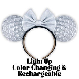 May include: Silver Minnie Mouse ear headband with a large bow and geometric design on the ears. The headband has the text "Light Up Color Changing & Rechargeable". The ears and bow are silver and the headband is black.