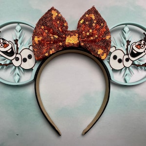 May include: Light blue Mickey Mouse ears with a snowflake design and Olaf the snowman. The ears are decorated with a large orange and gold bow.