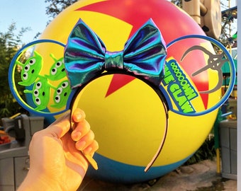 Puede incluir: Un par de orejas de Mickey Mouse azules y verdes con un lazo holográfico. Las orejas presentan un extraterrestre verde de Toy Story y el texto "Ooooooh Claw".