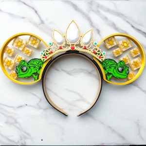 May include: A pair of yellow mouse ears with a green chameleon on each ear. The ears are decorated with a gold crown and a pattern of suns.