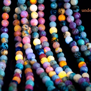 May include: Multiple strands of colorful gemstone beads glowing under a UV light. The beads are round and have a variety of colors, including pink, purple, blue, green, and yellow.
