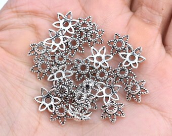 May include: A pile of silver-toned metal flower-shaped beads with a small hole in the center of each bead. The beads have a delicate, intricate design.
