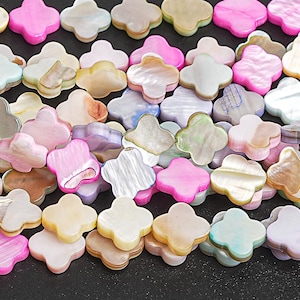 May include: Close-up of a collection of clover-shaped beads in various colors, including pink, white, and beige. The beads have a glossy, iridescent finish, and are arranged closely together. The beads are likely for jewelry making.