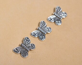 May include: Three silver butterfly charms with intricate details. The charms are arranged in a row on a light brown surface.