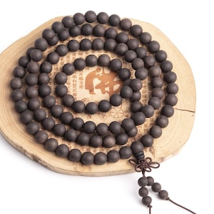 May include: A dark brown wooden mala bead necklace with a knotted tassel. The beads are round and smooth, and the necklace is coiled on a wooden base.