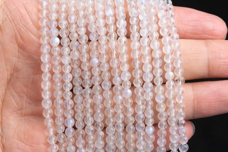 May include: A strand of white moonstone beads, each bead is round and smooth. The beads are strung on a clear thread and are arranged in a straight line. The beads are translucent and have a slight iridescent sheen.