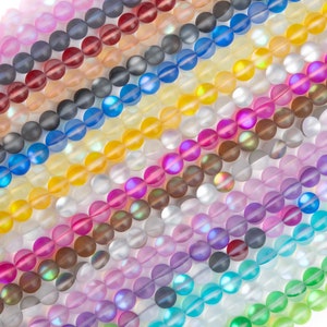 Puede incluir: Surtido de cuentas de vidrio iridiscente en varios colores, incluyendo rosa, azul, verde, amarillo y morado. Las cuentas son redondas y tienen un acabado liso y pulido.