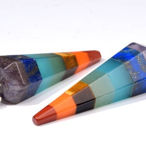 May include: Two faceted crystal pendulums with a rainbow color scheme. The crystals have a hexagonal shape and are arranged in a gradient of colors, including red, orange, yellow, green, blue, and purple. Each pendulum has a silver-colored metal loop.