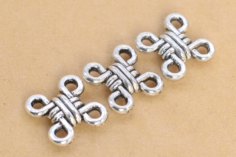 May include: Three silver metal decorative charms in the shape of a knot. The charms are intricately designed and have a polished finish.
