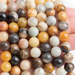 May include: A strand of round, smooth, multi-colored gemstone beads. The beads are a mix of brown, yellow, white, and black. The beads are strung on a clear string.