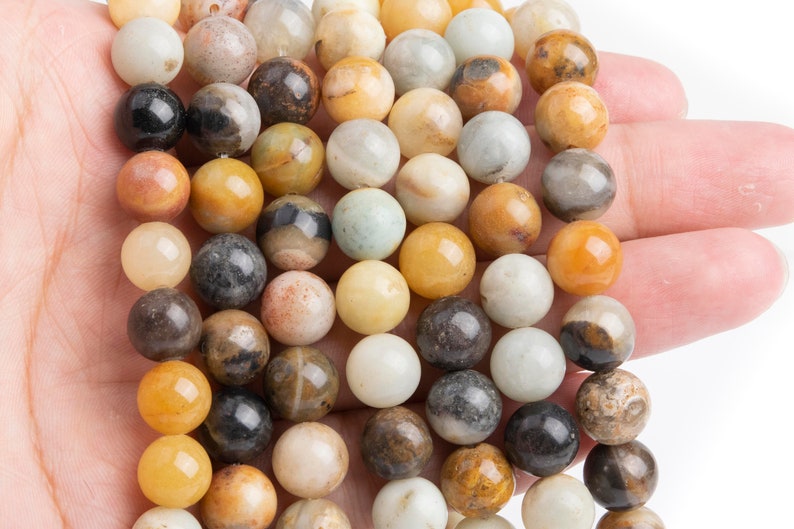 May include: A strand of round, smooth, multi-colored gemstone beads. The beads are a mix of brown, yellow, white, and black. The beads are strung on a clear string.