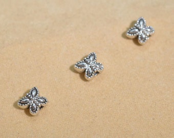 May include: Three small, silver-colored, butterfly-shaped beads are scattered on a light brown surface. Each bead has a detailed, textured design with a central element and four symmetrical wings. The beads appear to be made of metal.