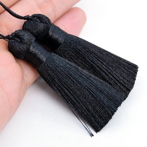 K&ouml;nnte beinhalten: Zwei schwarze Quastenb&auml;nder mit einem Knoten oben. Die Quasten bestehen aus d&uuml;nnen Stofff&auml;den und sind etwa 12,5 cm lang.