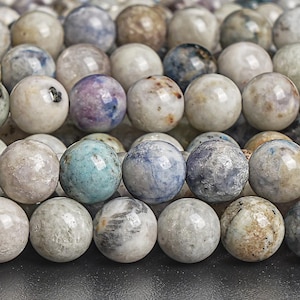 May include: A collection of round gemstone beads in various colors, including white, blue, purple, and tan. The beads have a polished, glossy finish and are arranged closely together, filling the frame. The natural stone beads are likely for jewelry making.