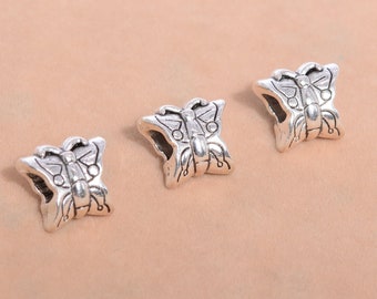 May include: Three silver-colored butterfly charms. Each charm has a detailed butterfly design with open wings and a textured surface. The charms are displayed on a light brown background, showcasing their intricate details.
