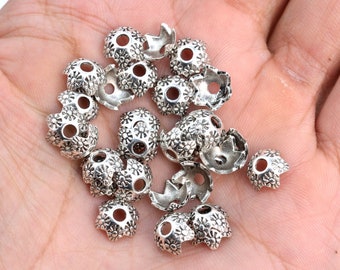 May include: A close-up of a pile of silver metal bead caps with a floral design. The bead caps have a hole in the center and are designed to be used for jewelry making.