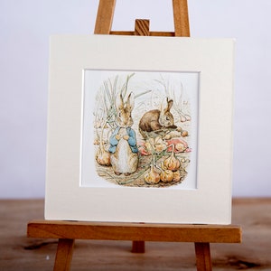 May include: A vintage illustration of two brown rabbits in a garden. The rabbit on the left is wearing a blue jacket and is standing upright. The rabbit on the right is lying down and is looking at the rabbit on the left. Both rabbits are surrounded by green plants and white onions.