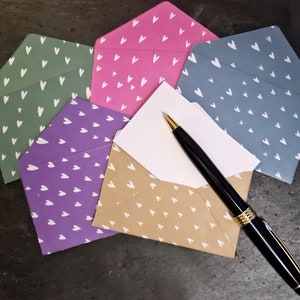May include: A collection of patterned envelopes in various colours: green, pink, purple, and blue, each with a white heart design. A black pen with gold detailing rests on an open envelope with a white card.
