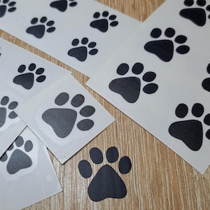Peut inclure: Feuilles de papier blanc avec des autocollants en forme de pattes de chien noires. Les empreintes de pattes sont de différentes tailles et sont disposées en rangées.