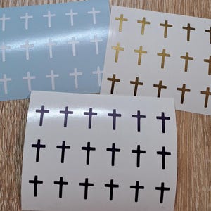 Puede incluir: Tres hojas de pegatinas con diseños de cruces. Una hoja es azul claro con cruces blancas, otra es blanca con cruces doradas y marrones, y la tercera es blanca con cruces negras. Las pegatinas están sobre una superficie de madera.