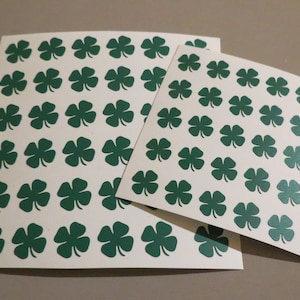 May include: Two sheets of white paper with green four-leaf clover stickers. The stickers are arranged in a grid pattern.