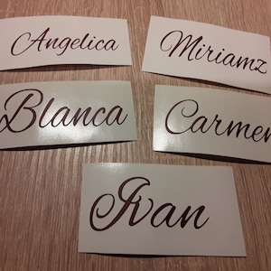 Puede incluir: Cinco pegatinas rectangulares con nombres en fuente cursiva. Los nombres son Angelica, Miriamz, Blanca, Carmen e Ivan. Los nombres son de color rojo oscuro, sobre una superficie de madera marrón claro.