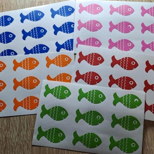 Puede incluir: Cuatro hojas de pegatinas con diferentes diseños de peces de colores. Los peces tienen todos la misma forma pero diferentes colores: azul, naranja, verde y rojo.