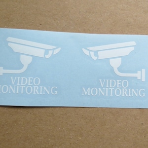 以下が含まれることがあります： 薄い青色の背景に、セキュリティカメラのシルエットと「VIDEO MONITORING」というテキストが書かれた、白いビニールステッカー2枚