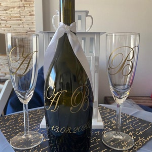 Puede incluir: Una botella de champán verde oscuro con las iniciales doradas "A B" y la fecha "13.08.2020", flanqueada por dos copas de champán. Las copas tienen las letras "A" y "B" en dorado. Una cinta blanca adorna la botella.