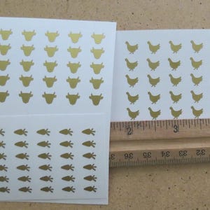 Op de afbeelding: Drie vellen witte stickers met gouden folie boerderijdierontwerpen. De stickers bevatten koeien, kippen en wortelen. Een houten liniaal ligt naast de stickers en meet in centimeters.