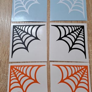 Op de afbeelding: Zes vierkante stickers met een spinnenwebontwerp. Twee zijn lichtblauw met witte webben, twee zijn wit met zwarte webben en twee zijn wit met oranje webben. De stickers zijn gerangschikt op een lichtbruin oppervlak.