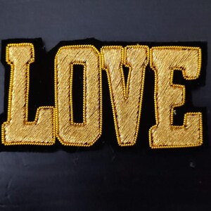 Peut inclure: Un patch brodé or avec le mot "LOVE" en grandes lettres majuscules sur un fond de velours noir.