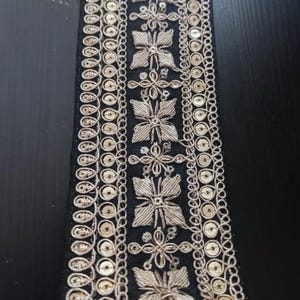 以下が含まれることがあります： 黒い生地のトリムに金色の刺繍とスパンコールが施されています。トリムは、花柄のデザインが繰り返されるパターンになっています。