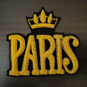 Peut inclure: Un patch brodé noir et or avec le mot "PARIS" dans une police de caractères stylisée. Le mot est entouré d'une couronne dorée.