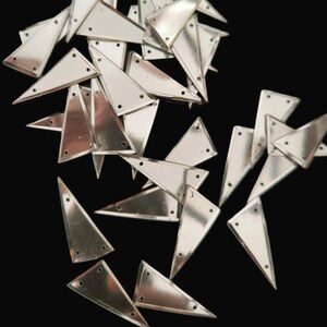 Pode incluir: Uma coleção de enfeites triangulares de metal prateado, cada um com três pequenos orifícios. Os triângulos estão espalhados sobre um fundo preto, refletindo a luz. Estas peças brilhantes são provavelmente para artesanato ou moda.