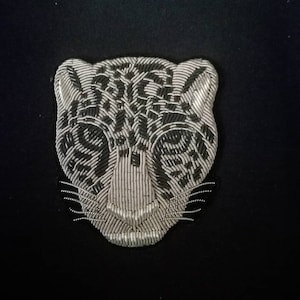 Peut inclure: Gros plan d'une tête de léopard perlée argentée sur fond noir. Le visage du léopard est méticuleusement réalisé avec des perles argentées, soulignant ses traits. Ce motif pourrait être une application ou un écusson pour vêtements ou accessoires.