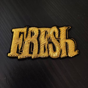 Peut inclure: Un patch brodé or avec le mot "FASH" dans une police de caractères stylisée.