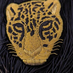 Könnte beinhalten: Ein gold gestickter Jaguar-Kopf-Patch mit schwarzen Details. Der Patch ist auf einem schwarzen Hintergrund.