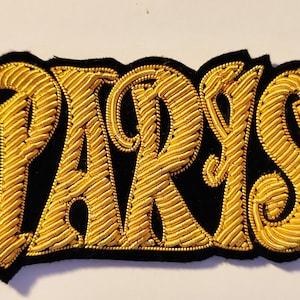 Könnte beinhalten: Gold gestickter "PARIS" Patch auf schwarzem Filzgrund.