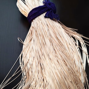 Peut inclure: Un faisceau de fins fils de couleur or clair, solidement liés par une corde violet foncé. La corde est nouée en haut, fixant les fils. Les fils s'évasent en bas, créant un effet texturé.
