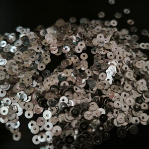 Peut inclure: Un tas de paillettes argentées, plates et rondes, avec un trou au centre. Les paillettes sont dispersées sur une surface sombre, créant un aspect texturé et réfléchissant. Idéal pour les travaux manuels.