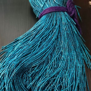 Pode incluir: Um pacote de contas de vidro azul turquesa, amarradas com uma fita roxa. As contas são pequenas e têm um acabamento brilhante e metálico.