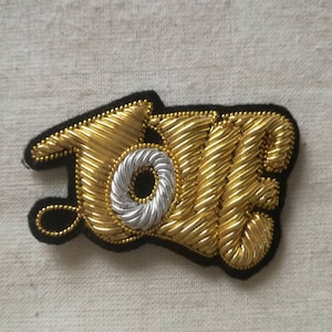 Puede incluir: Un parche bordado en oro y plata con la palabra "LOVE" en una fuente estilizada.