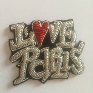 Può includere: Una toppa ricamata in argento con le parole "LOVE PARIS" in un font stilizzato. Un cuore rosso è al centro della toppa.