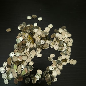 Op de afbeelding: Een stapel kleine, metallic goudkleurige ovale pailletten met twee gaatjes aan de bovenkant. De pailletten zijn verspreid op een donkere ondergrond. Ideaal voor knutselprojecten.