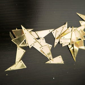 Pode incluir: Vista aérea de peças geométricas de metal dourado sobre uma superfície escura. As peças são triangulares e retangulares, com pequenos orifícios. A disposição sugere uma forma animal estilizada. Medidas métricas.
