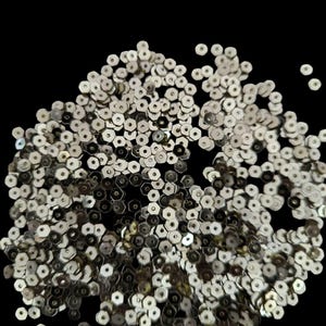 May include: A collection of silver sequins with a hexagonal shape, scattered against a black background. The sequins have a metallic sheen, reflecting light. Ideal for embellishing clothing, accessories, or craft projects.